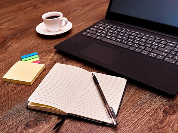 laptop on a desk with notebook, pen, colourful sticky note pads and a cup of coffee
